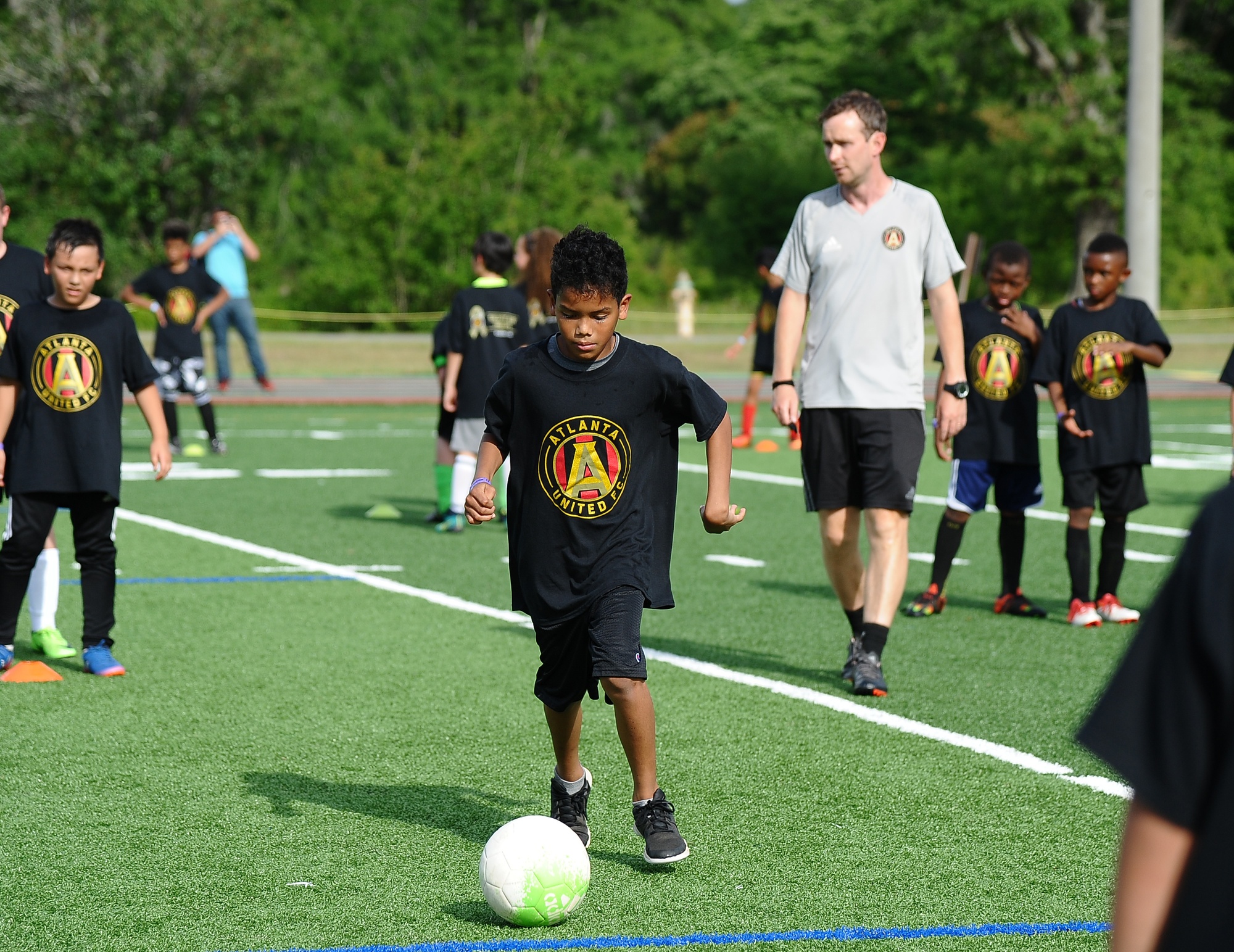 Child controlling a football on an outdoor pitch while an adult coach observes the practice