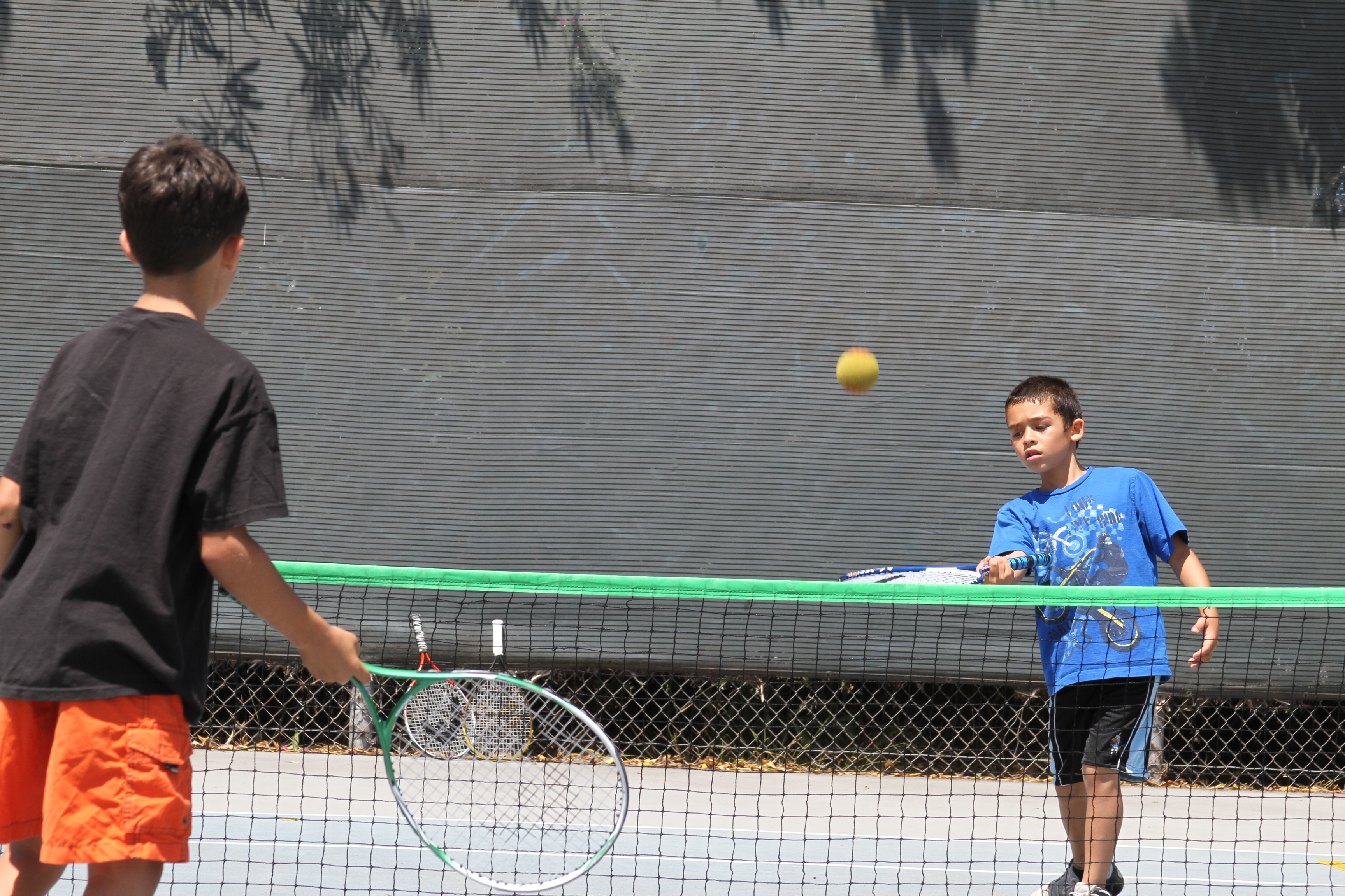 Two children using rackets and a slower ball across a low net during a supervised tennis clinic