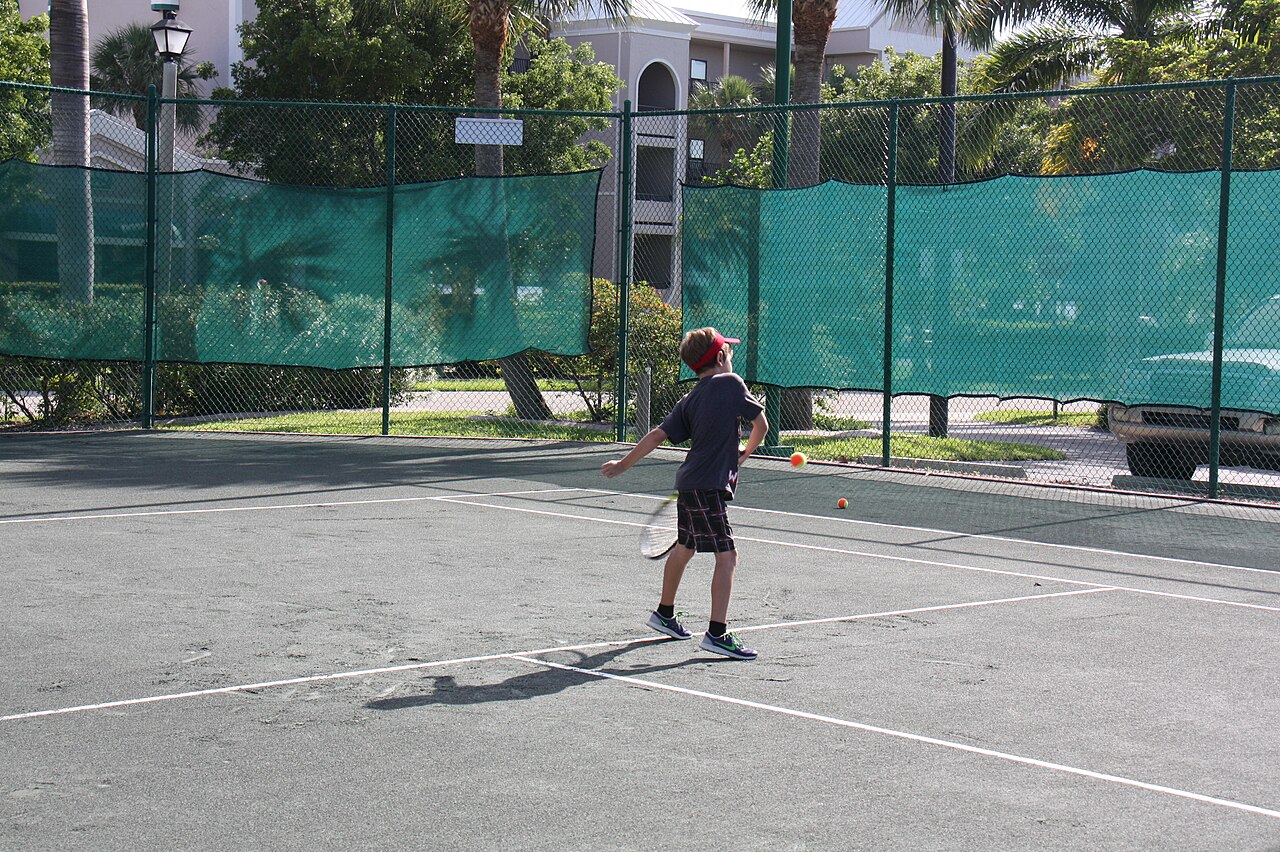 Child holding a tennis racket while preparing the next ball during an outdoor tennis camp