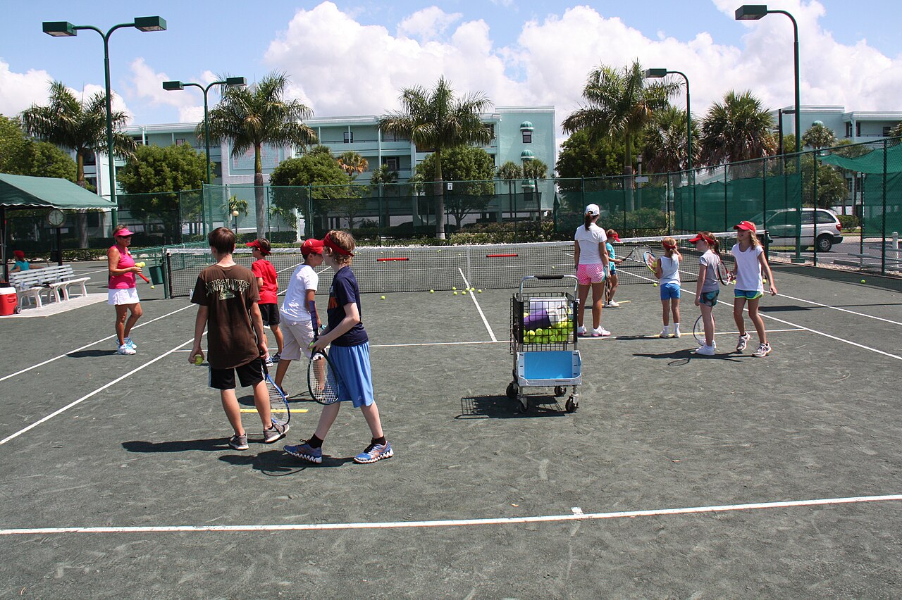 Children holding tennis rackets beside adult leaders and a ball cart during an outdoor tennis camp